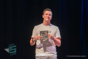 Ein weiterer Gast auf der Bühne der Lesung von Laura Purcell im Theater am Ring in Saarlouis, der ein Buch präsentiert